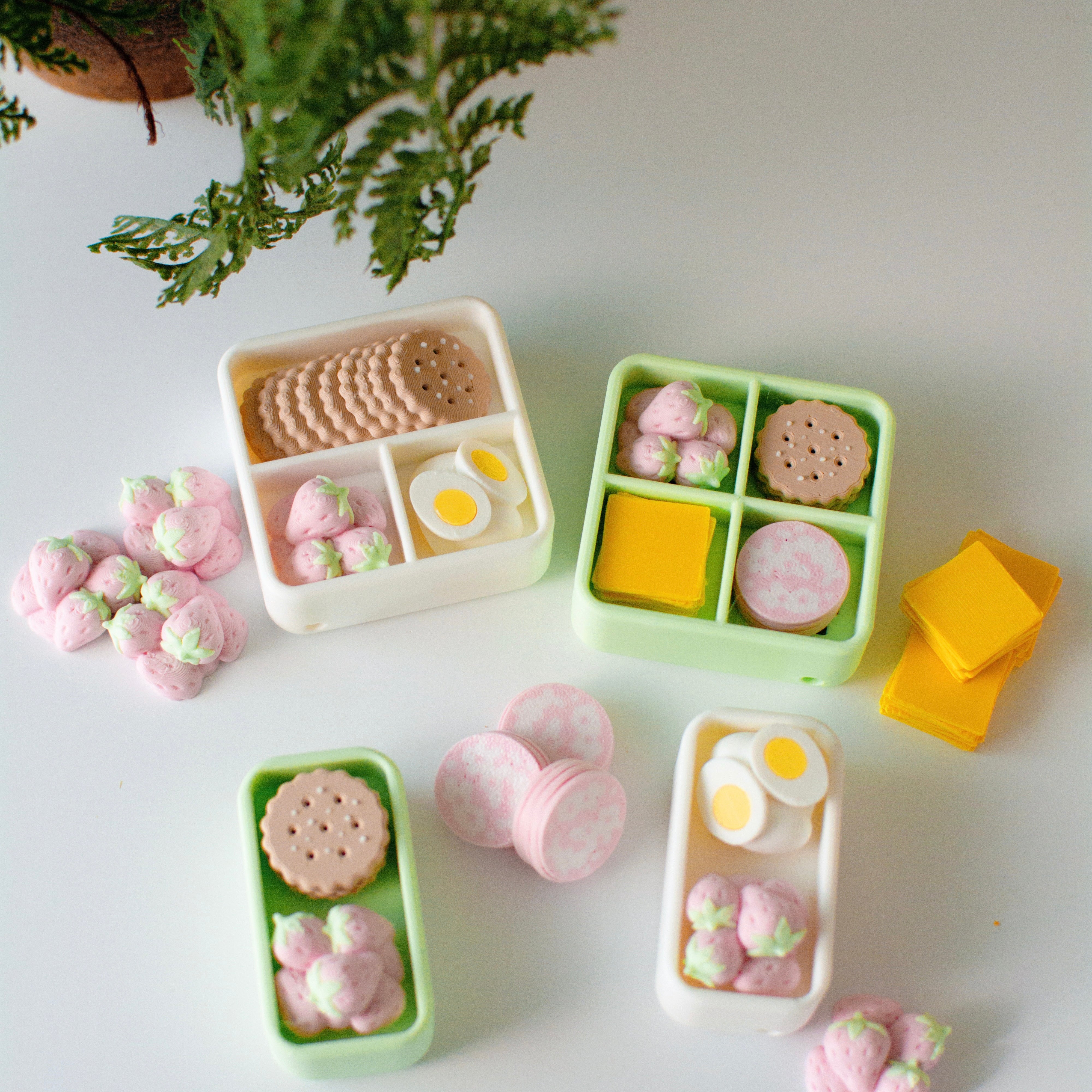 Miniature bento boxes with food items on a light surface with a plant in the corner.