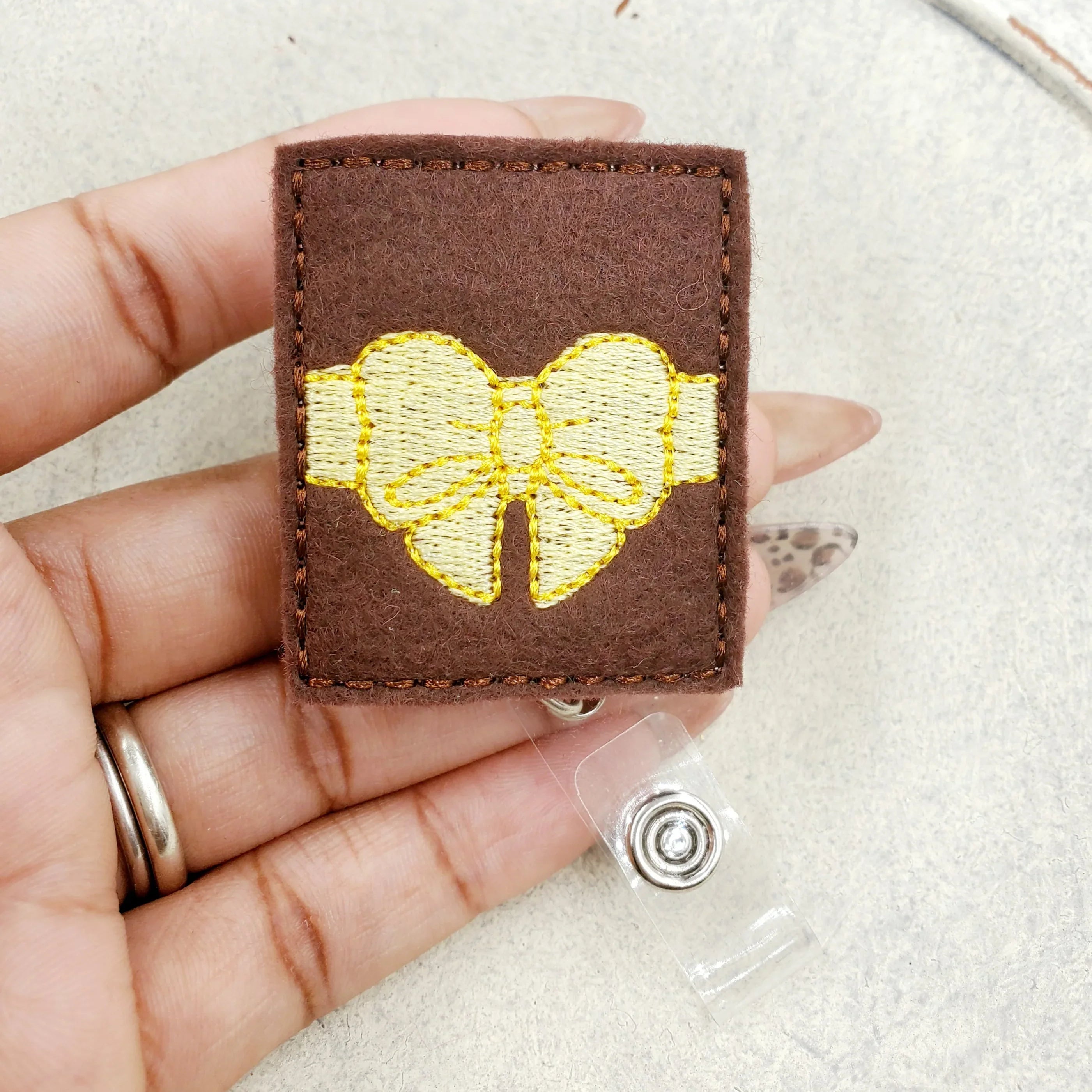 Hand holding a brown felt badge reel with yellow embroidered bow design