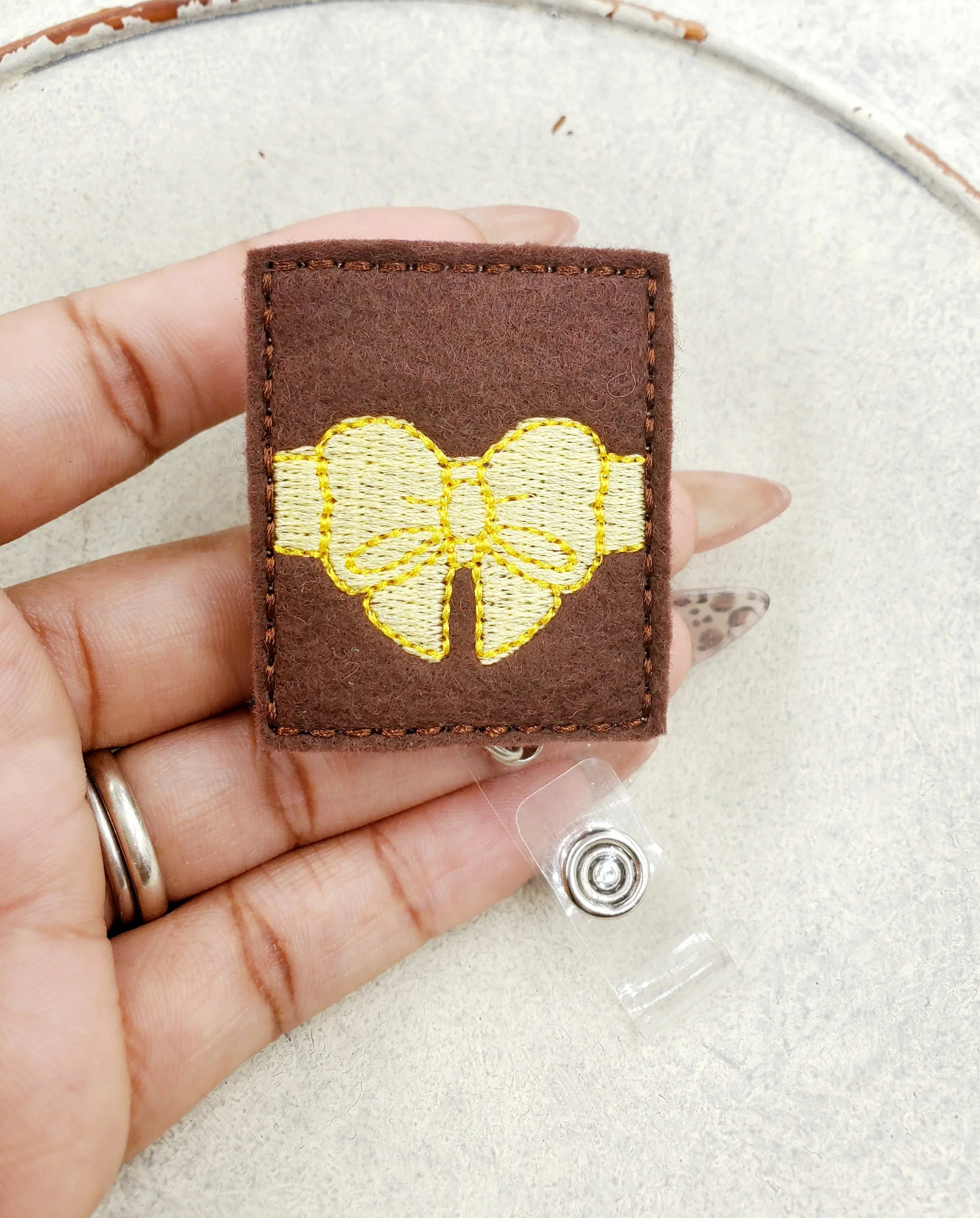 Hand holding a brown felt badge reel with yellow embroidered bow design