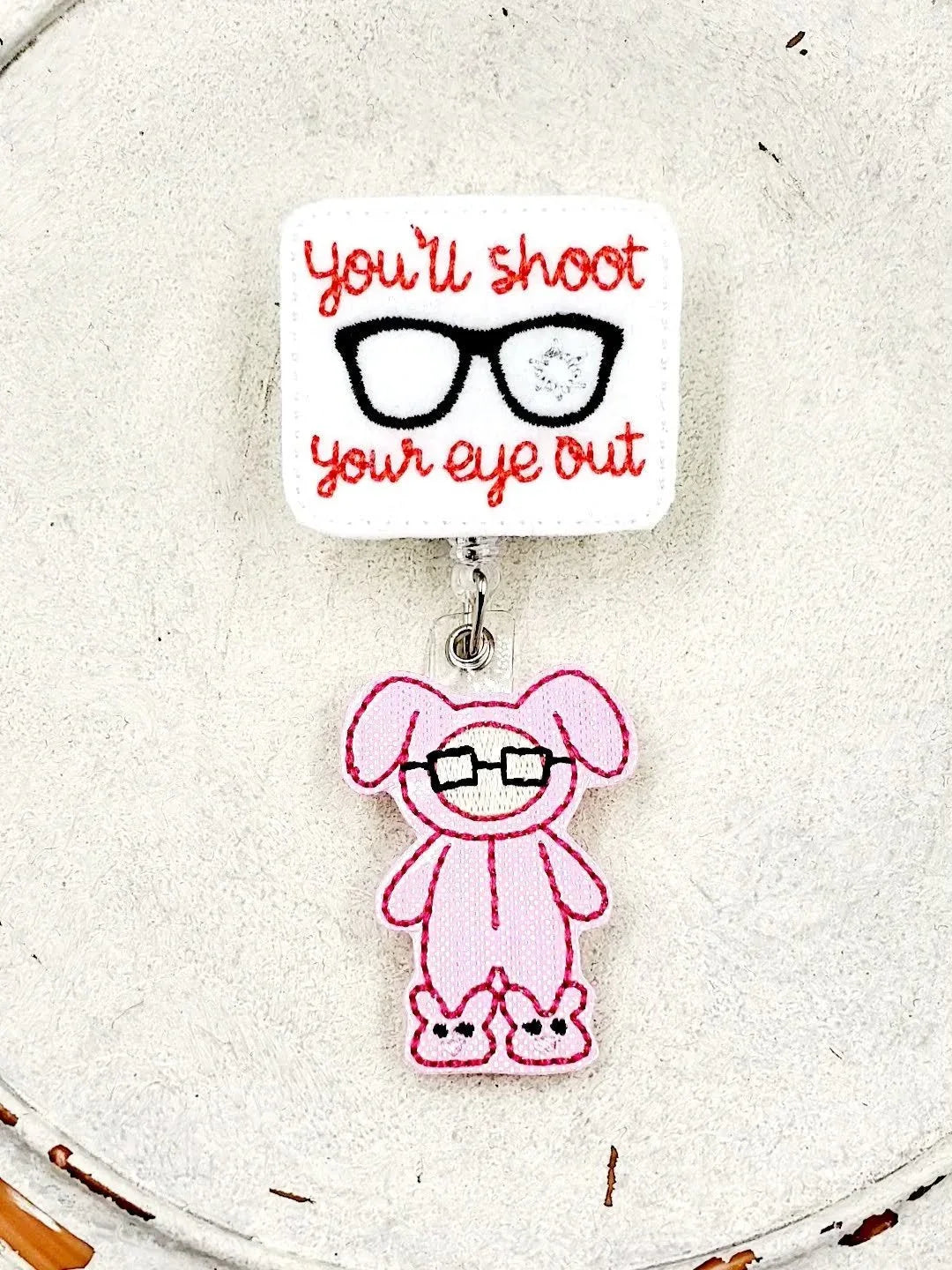 Embroidered badge reel with 'you'll shoot your eye out' text and pink bunny glasses charm.