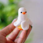 Hand holding a small white duck banana figurine with orange beak and feet against a blurred green and white background