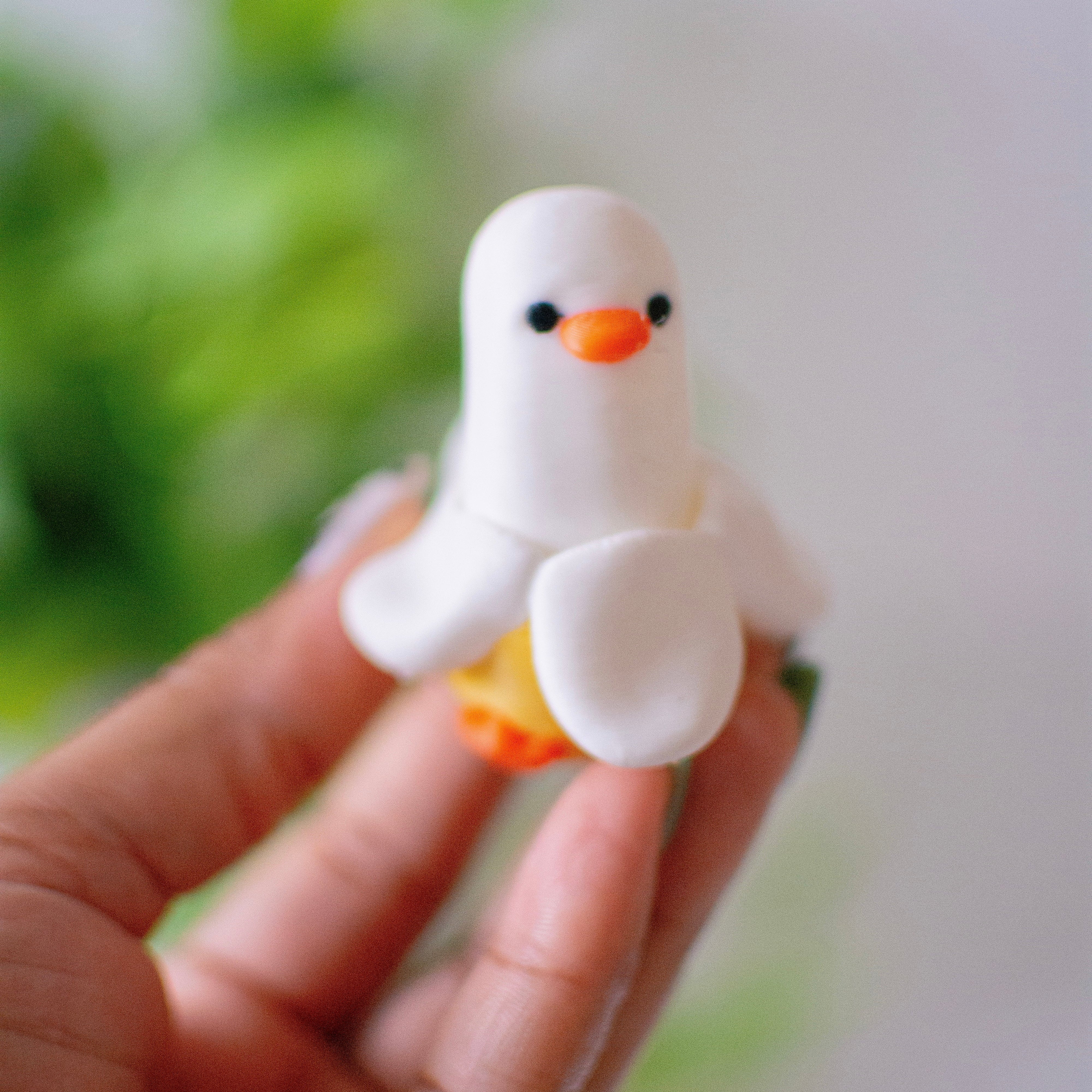 Hand holding a small white duck banana figurine with orange beak and feet against a blurred green and white background