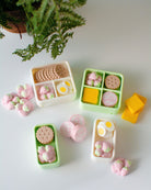 Miniature bento boxes with food items on a light surface with a plant in the corner.