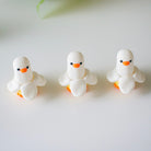 Three small white duck figurines with orange beaks on a light surface.
